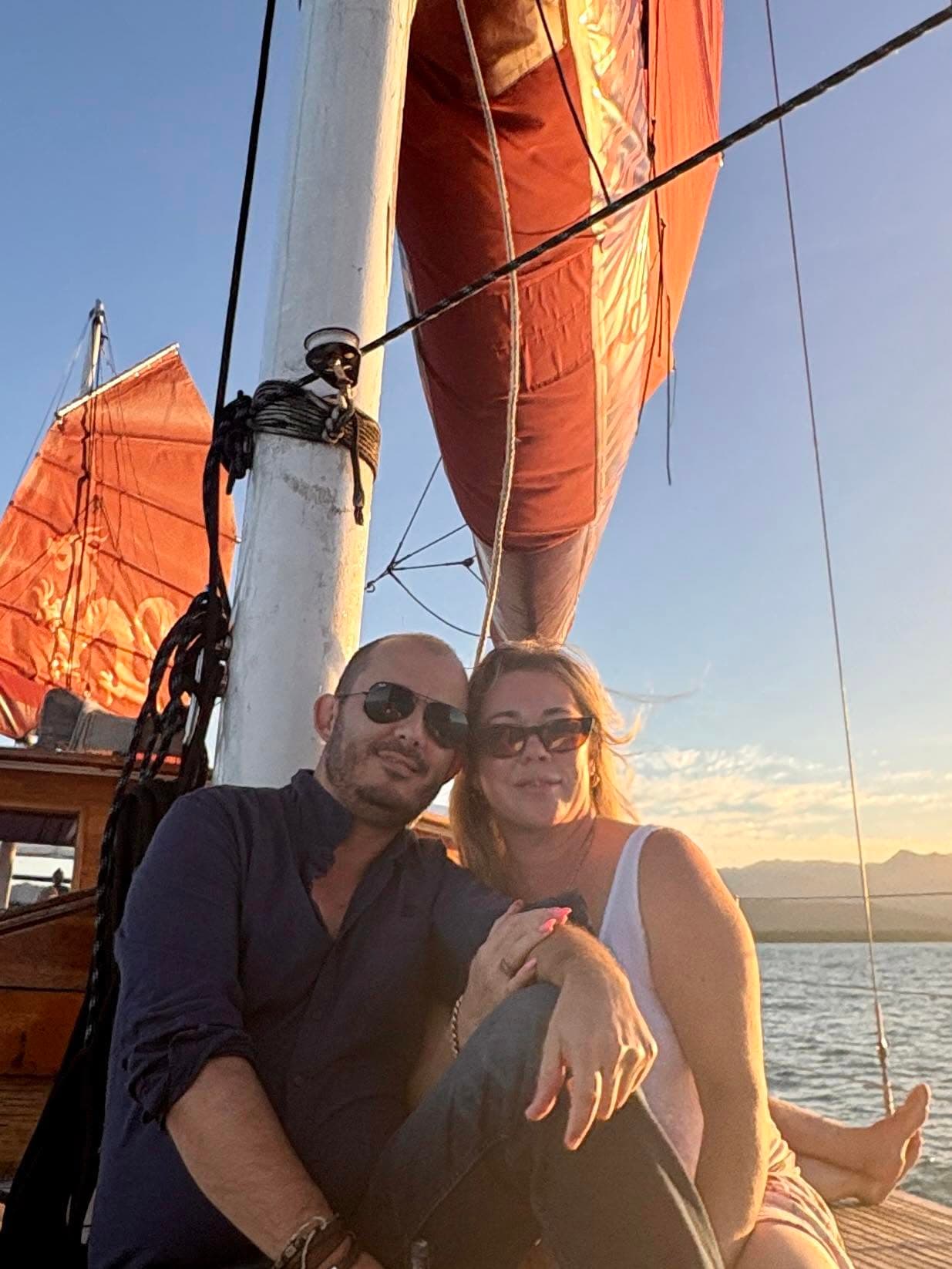 Two people aboard a sailing boat beneath orange sails in the evening light